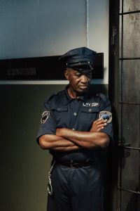 A police officer standing indoors in uniform, arms crossed, beside a door.