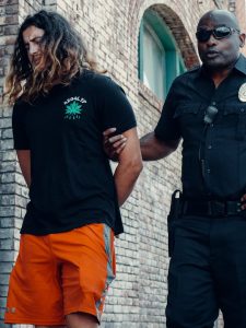 Police officer escorting a suspect with long hair along a brick wall, showcasing law enforcement in action.